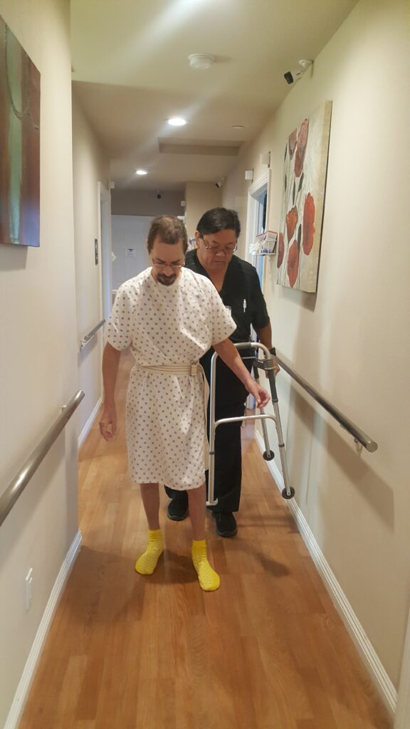 A man in a hospital gown and nurse are walking down the hallway.